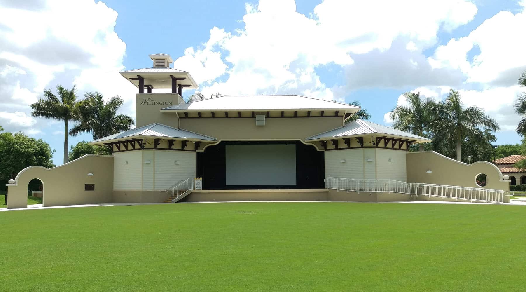 The Wellington Amphitheater outdoor performance venue with a covered stage, large projection screen, green lawn seating area, and palm trees under a partly cloudy sky in Wellington, Florida.