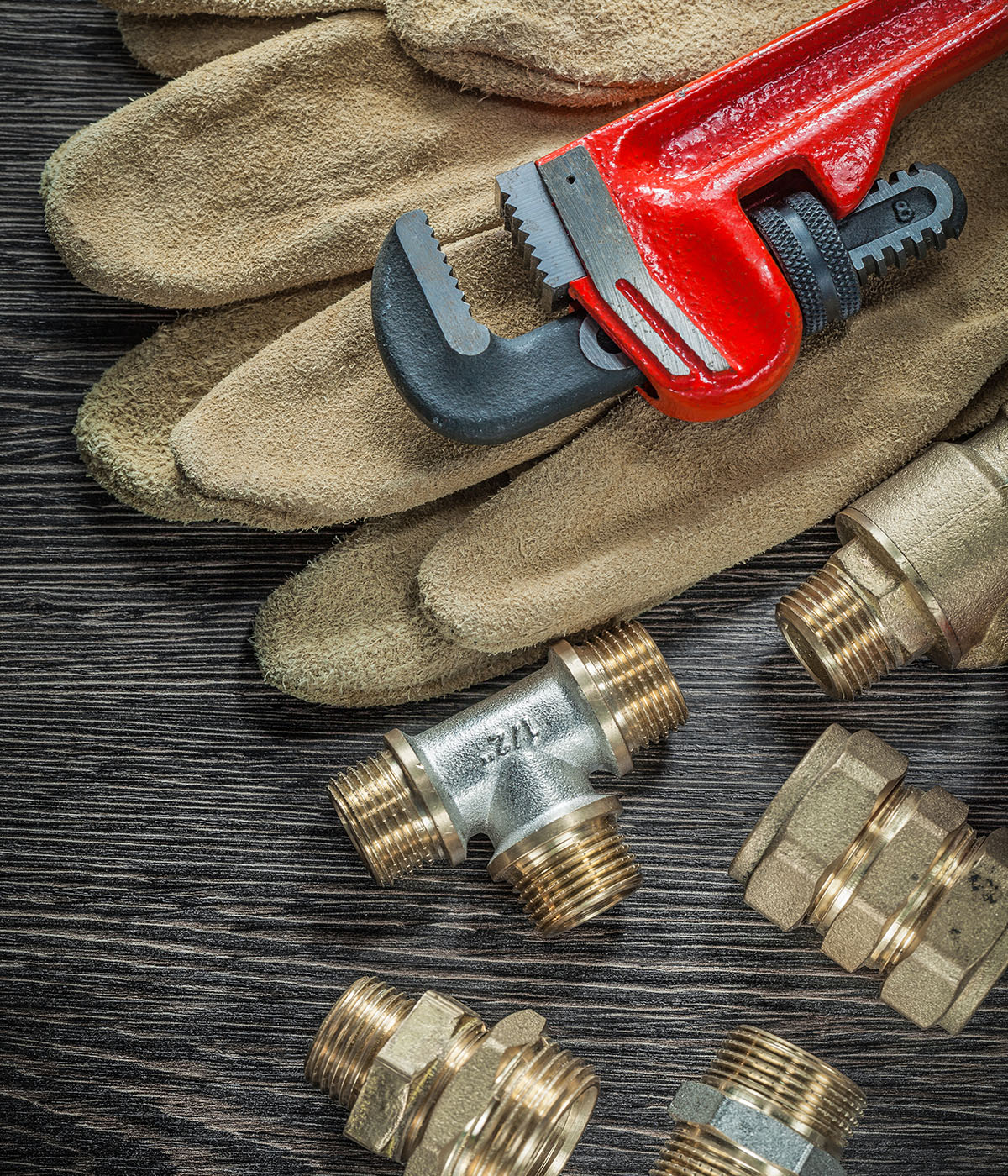 Assorted brass pipe fittings and a red pipe wrench laid out on tan leather work gloves against a dark wood surface