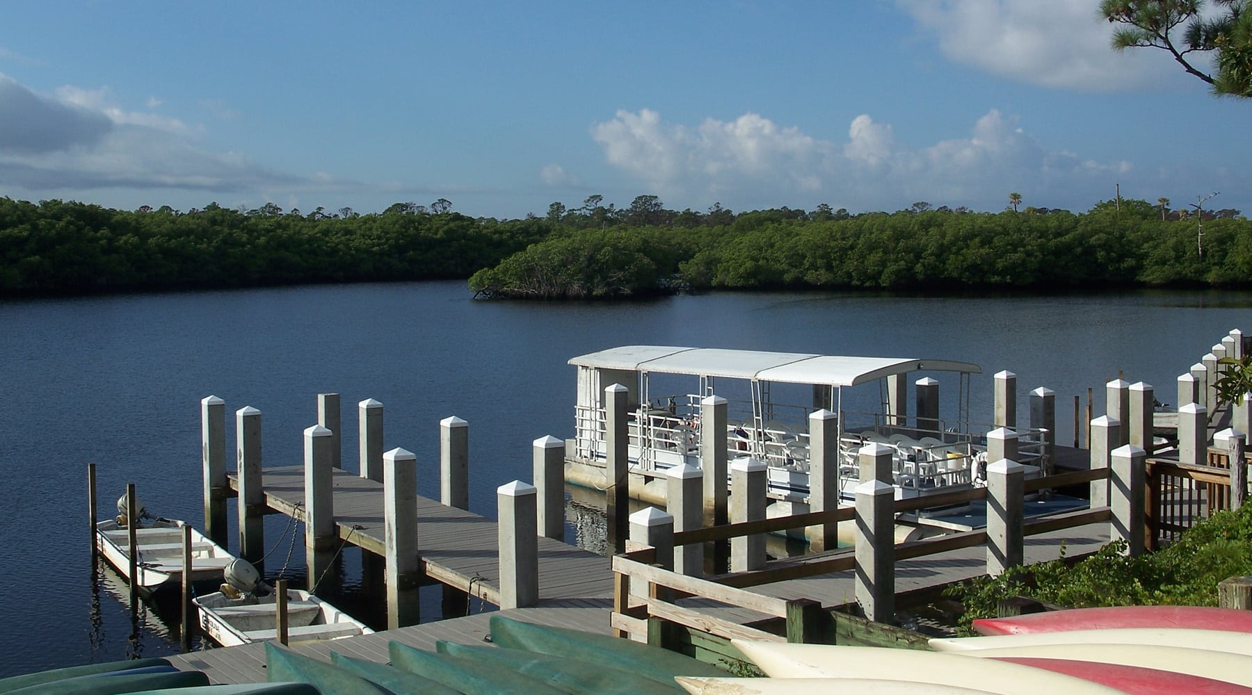 A tranquil waterway dock in Hobe Sound, Florida with a covered pontoon boat, wooden pier, small motorboat, colorful kayaks, and lush mangrove trees lining the shoreline under a partly cloudy sky.