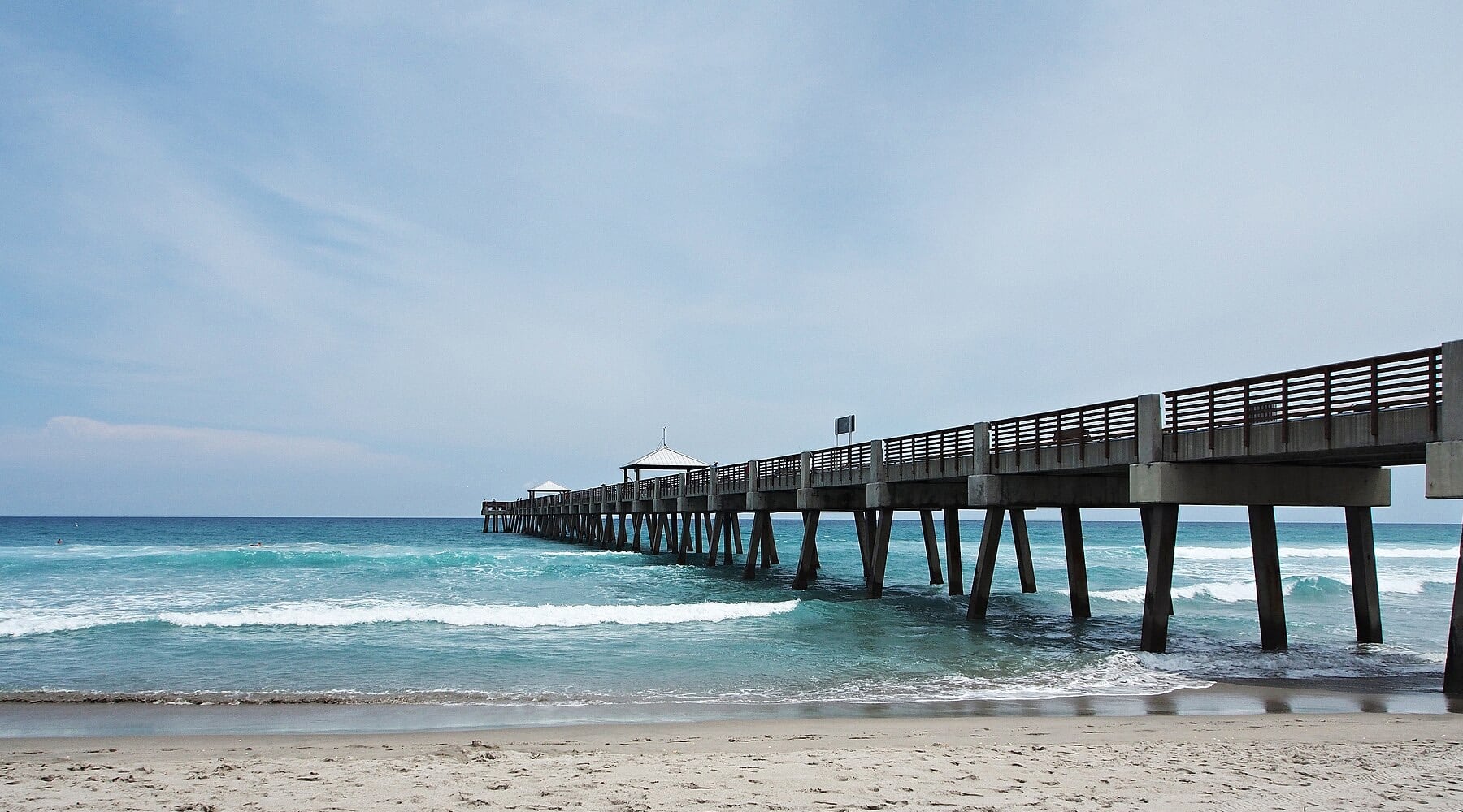 The Juno Beach Pier stretching over turquoise Atlantic Ocean waves with a white sandy beach in the foreground under a soft cloudy sky in Juno Beach, Florida.