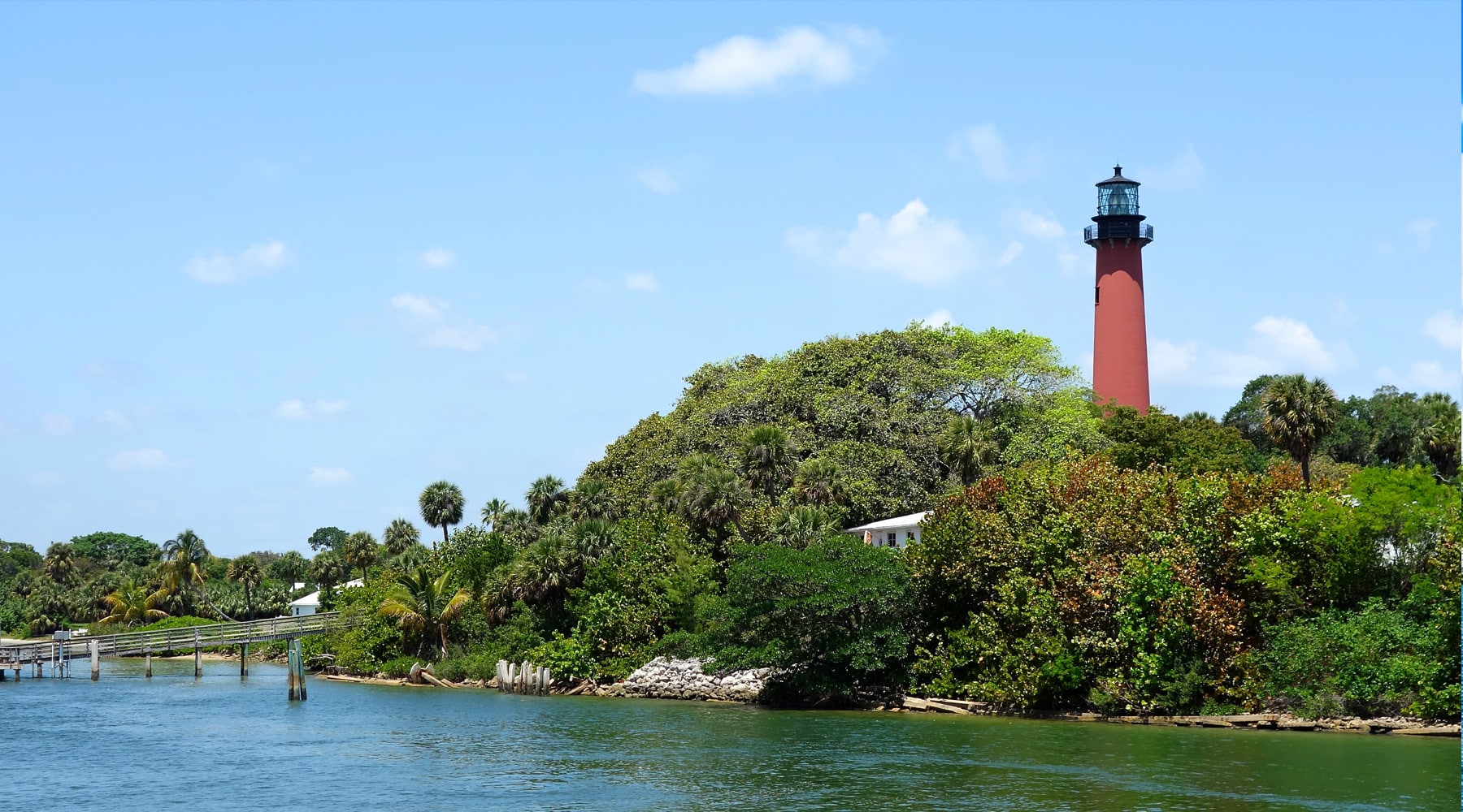 Scenic view of the Jupiter Lighthouse, a tall red brick tower rising above lush tropical trees and palm trees along the Loxahatchee River in Jupiter, Florida