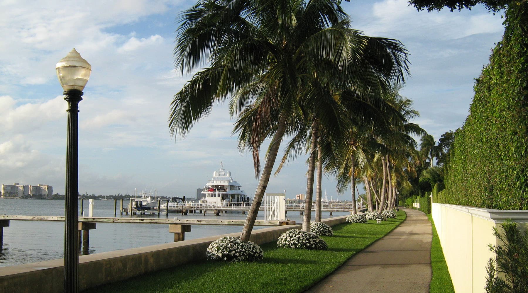 scenic waterfront walkway in Palm Beach, Florida lined with tall palm trees, manicured hedges, and white flower beds along the Intracoastal Waterway with a luxury yacht docked in the background.