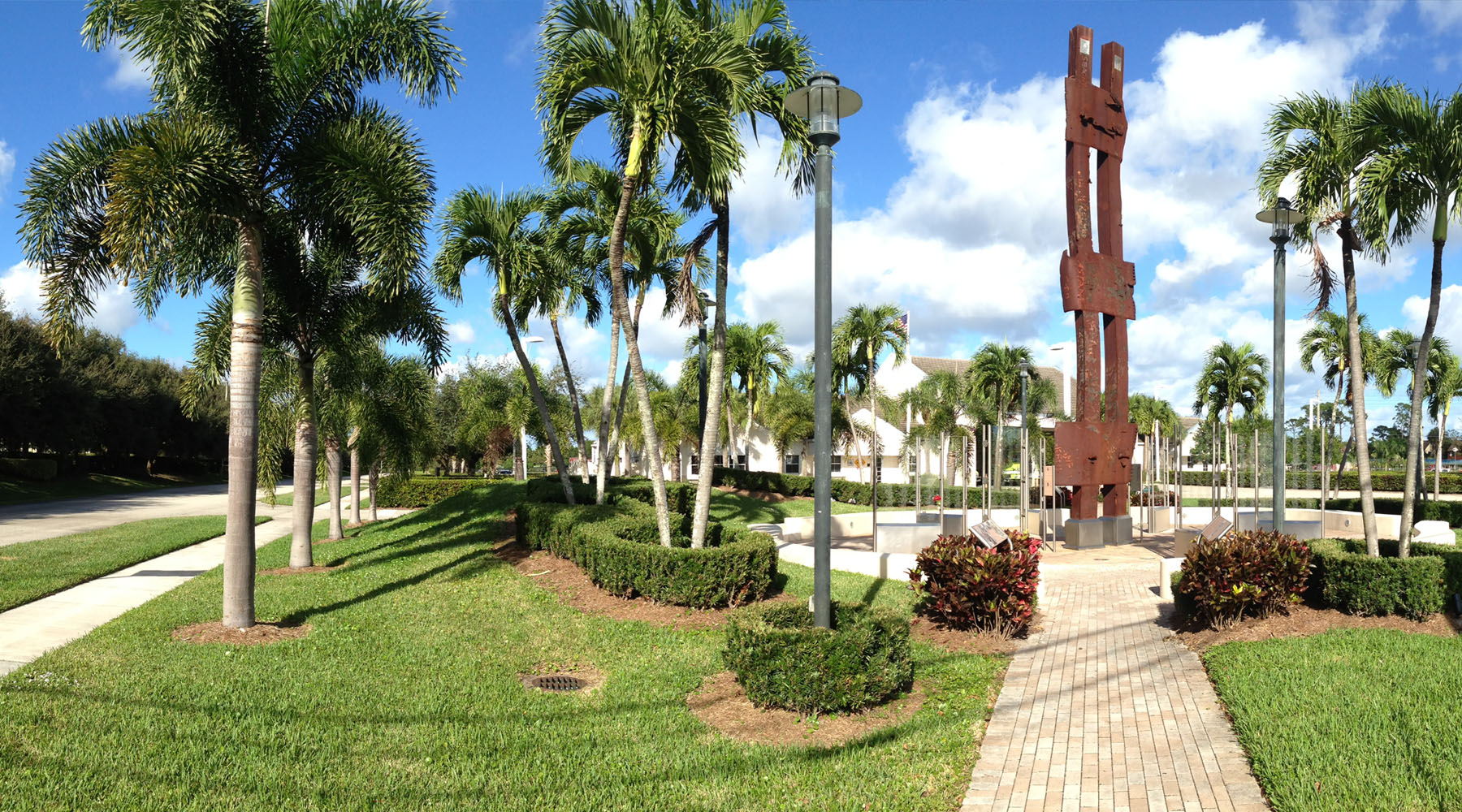 A public park in Palm Beach Gardens, Florida featuring a tall rusted steel memorial sculpture, brick walkway, manicured landscaping, and royal palm trees under a bright blue sky.