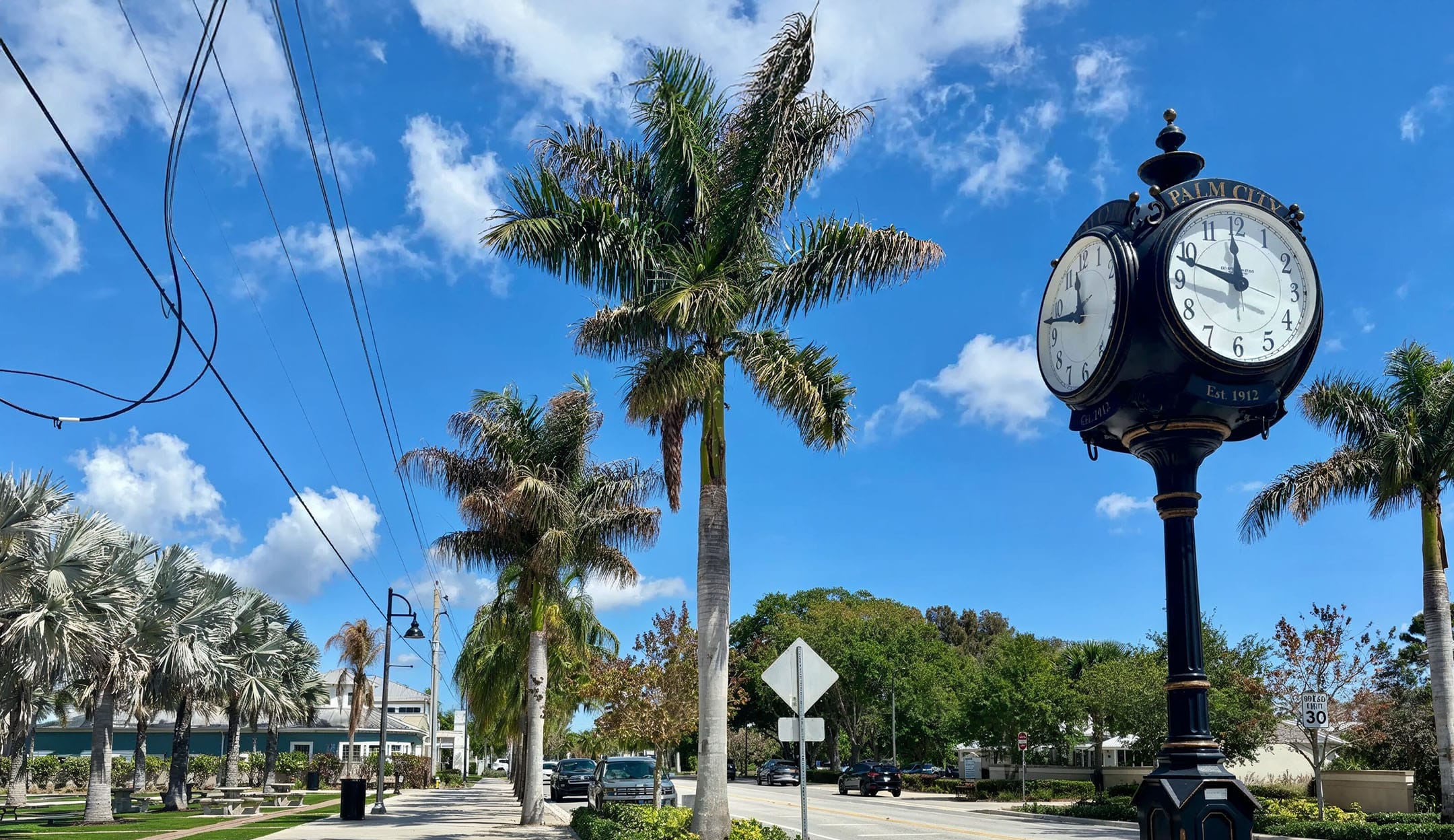 A sunny street scene in Palm City, Florida featuring a black and gold town clock established in 1912, palm tree-lined roads, and a bright blue sky with scattered clouds