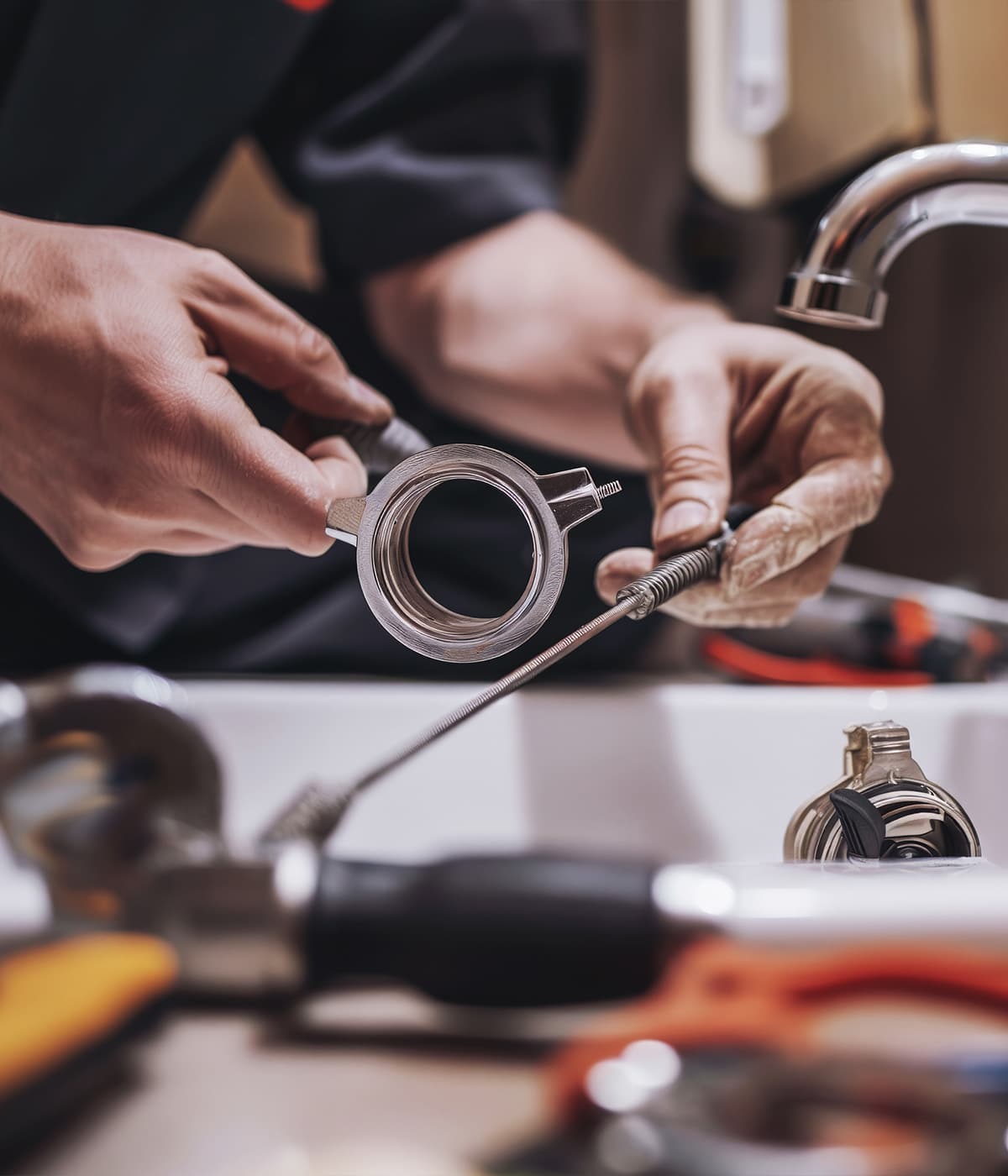 A plumber in a dark uniform using a drain snake and wrench to repair a bathroom sink faucet, with plumbing tools scattered on the sink in the foreground