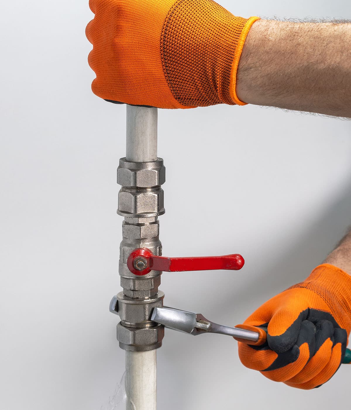 A plumber wearing orange work gloves using a wrench to tighten a silver ball valve with a red handle on a water supply pipe against a white background.