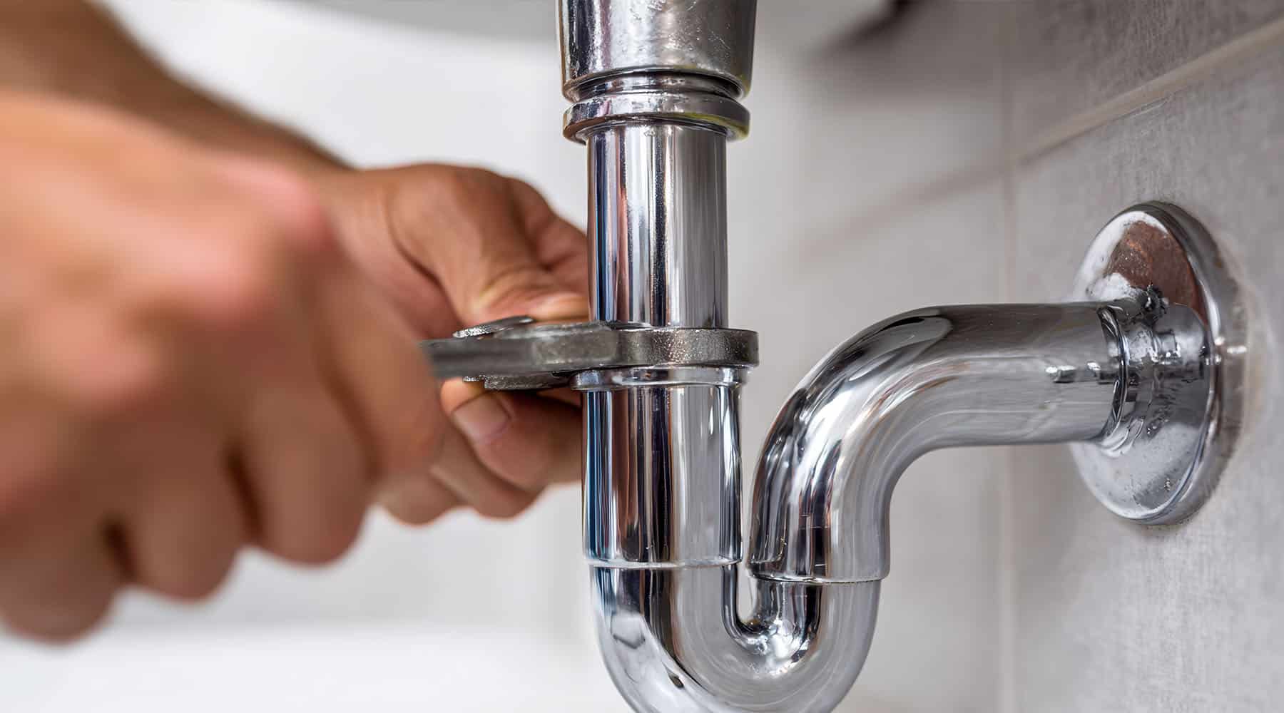 A close-up of a plumber's hand tightening the slip nut on a chrome bathroom sink P-trap drain pipe against a white tiled wall.