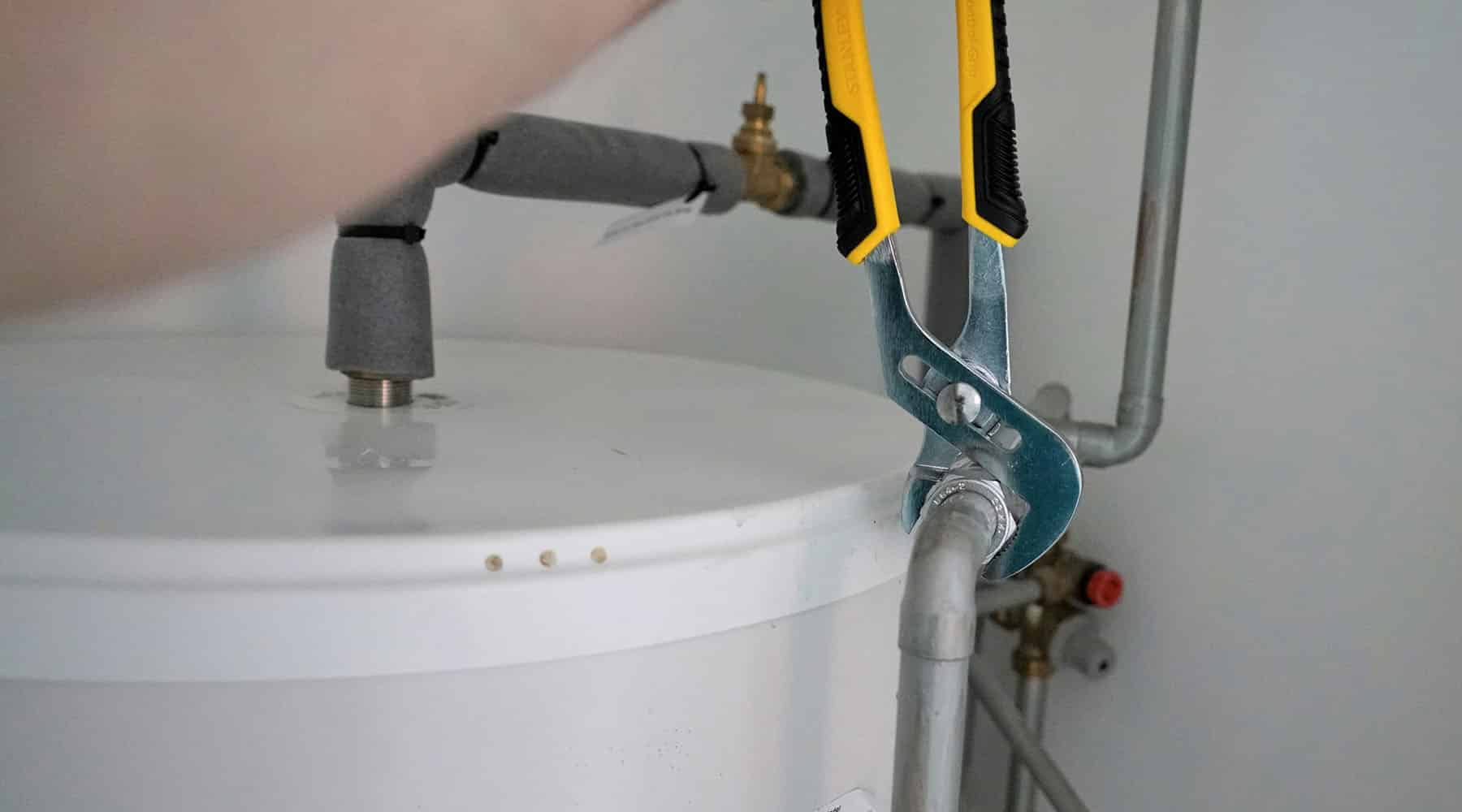 A plumber using yellow-handled pliers to tighten a pipe fitting on top of a white residential water heater tank with insulated supply lines and a brass shut-off valve.