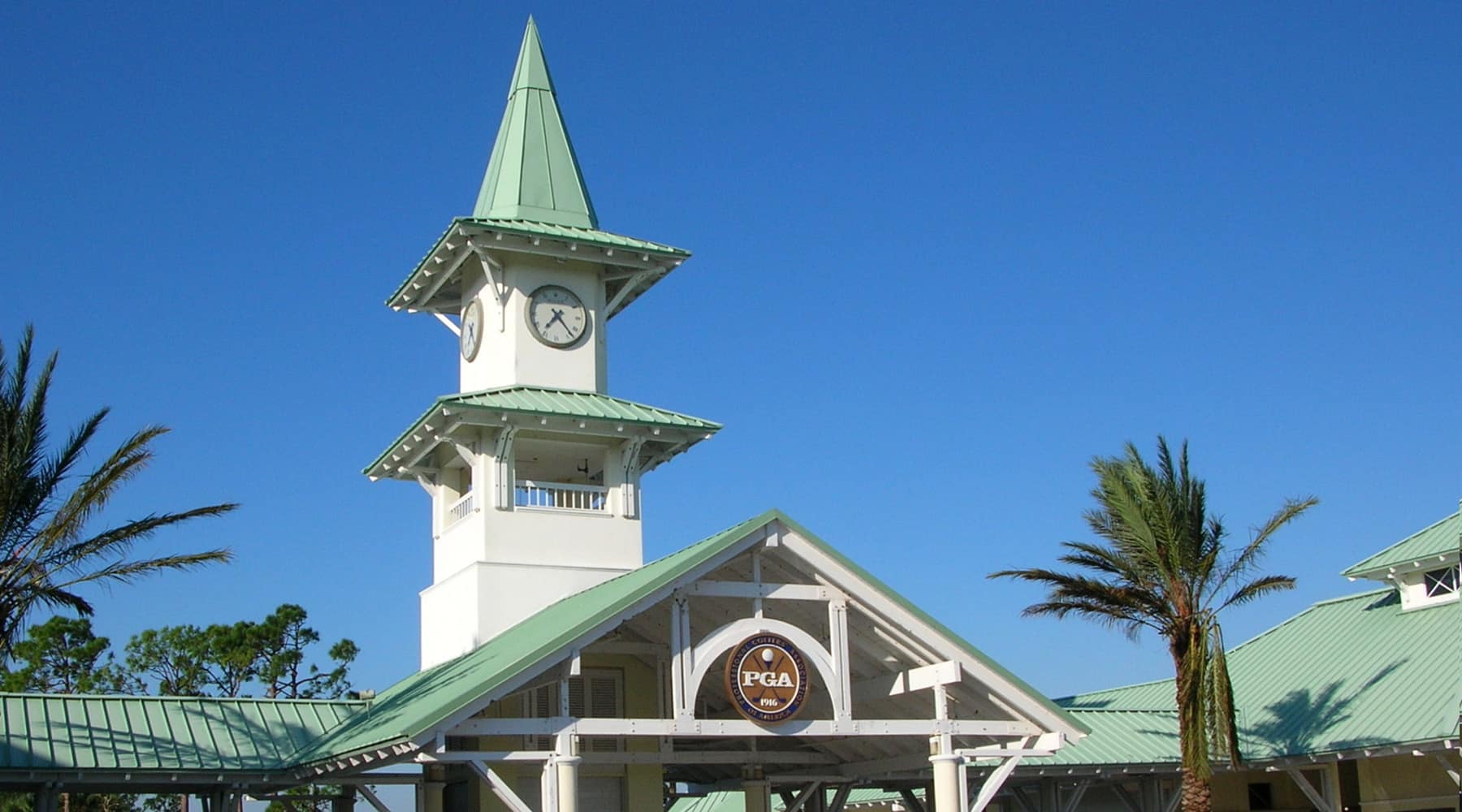 The entrance to PGA Village in Port St. Lucie, Florida, featuring a white clock tower with a green metal roof, the PGA of America seal, and palm trees under a clear blue sky.