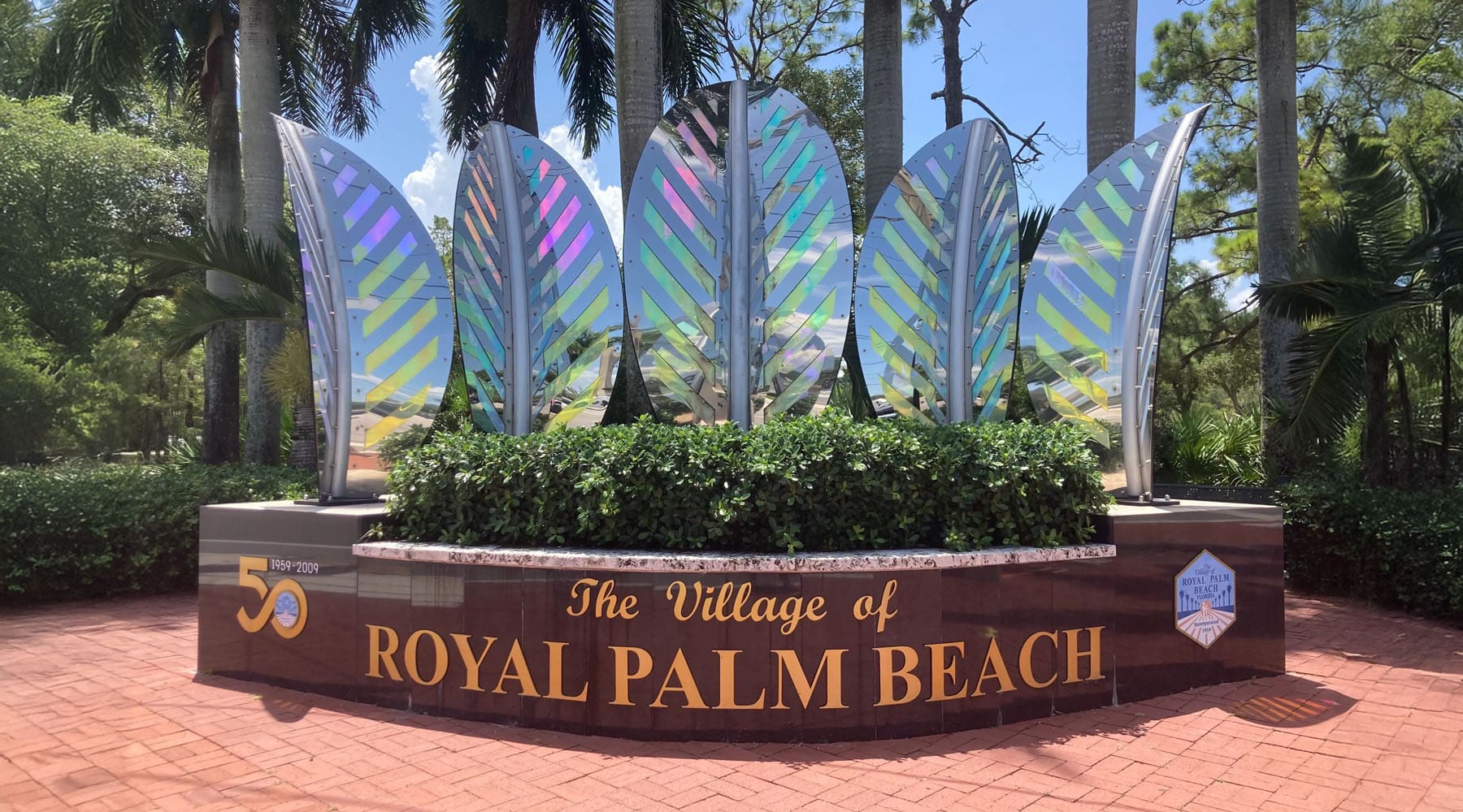 The Village of Royal Palm Beach entrance monument featuring iridescent metallic palm leaf sculptures on a decorative base surrounded by lush greenery and palm trees in Royal Palm Beach, Florida.