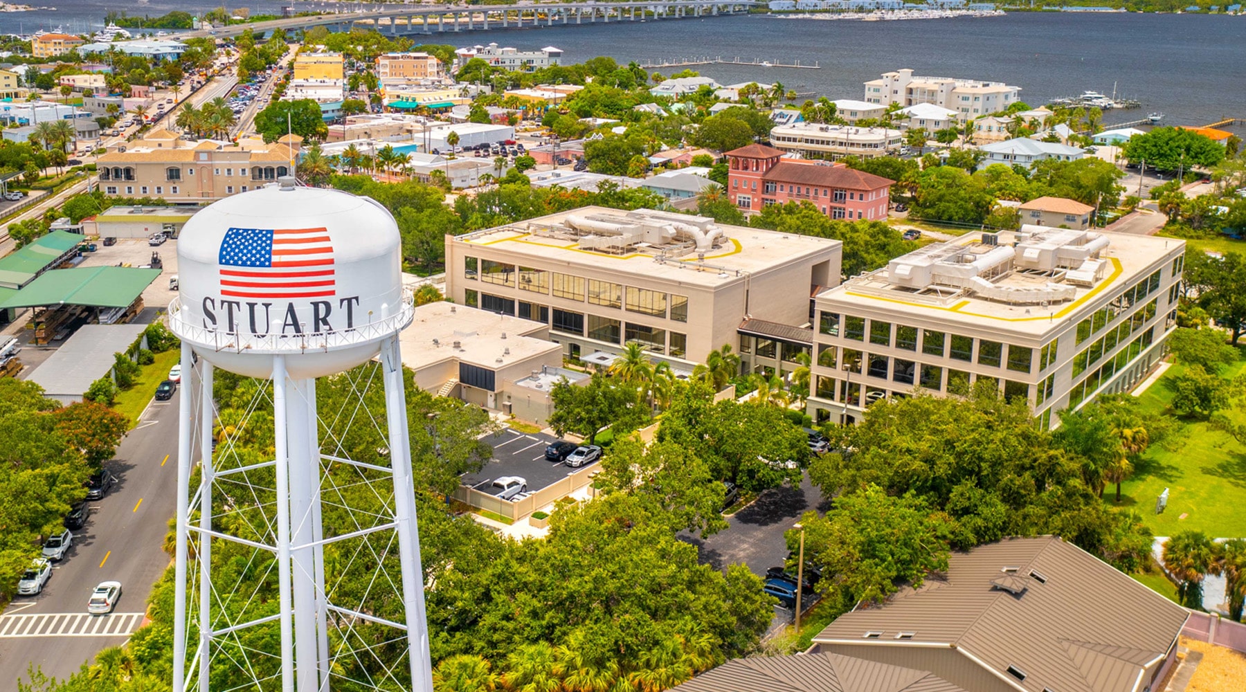 Aerial view of Stuart, Florida featuring the iconic white water tower with an American flag, surrounded by downtown buildings, palm trees, and the St. Lucie River in the background at sunset.
