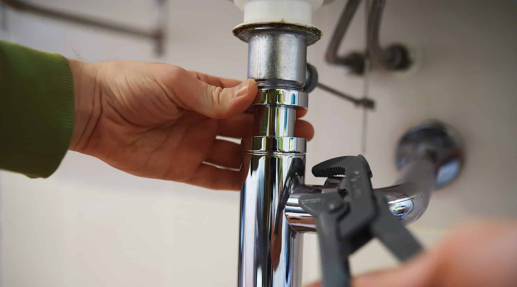 A close-up of a hand tightening the slip nut on a chrome sink drain pipe under a cabinet while a pipe wrench holds the fitting in place.