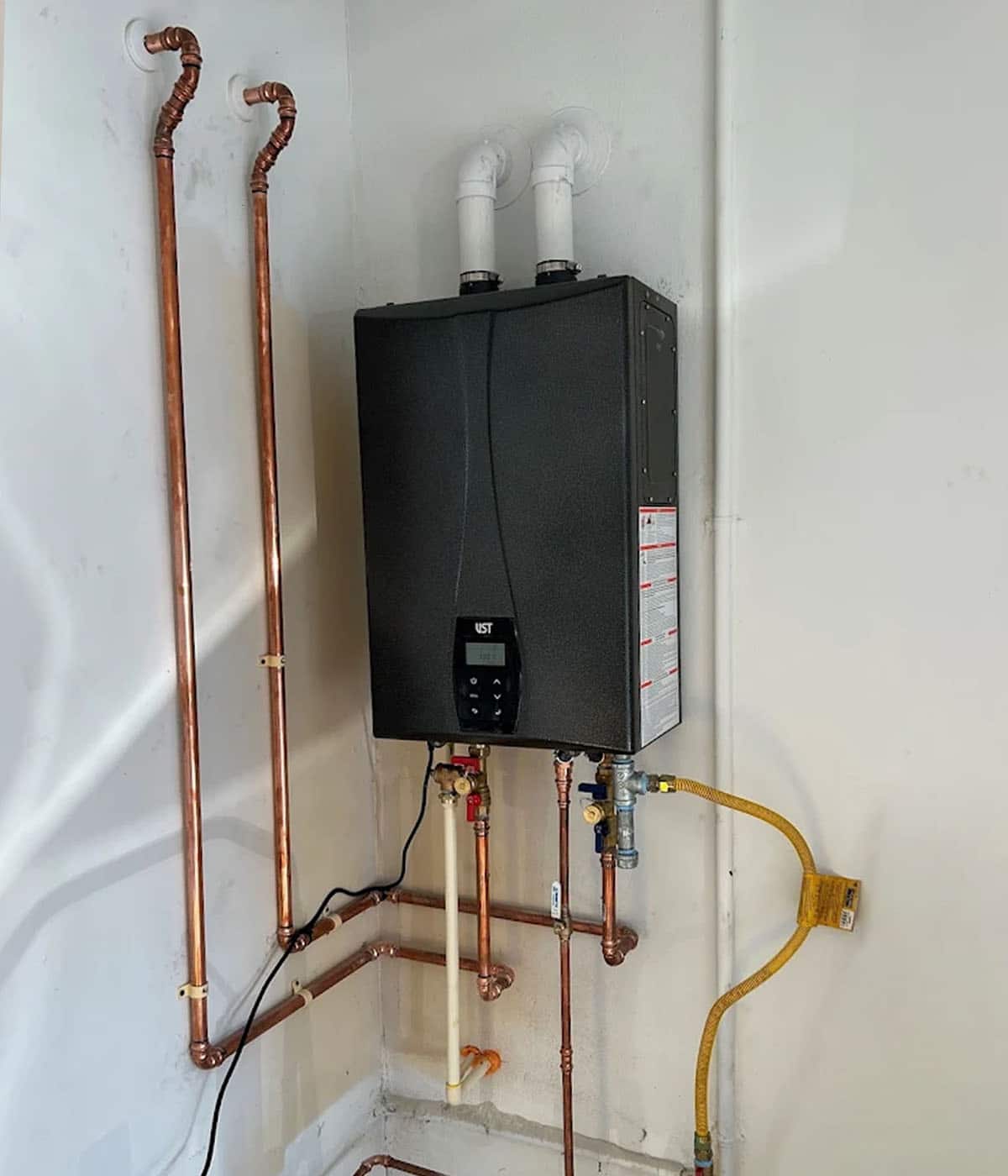 A wall-mounted tankless water heater with copper pipe connections, white PVC exhaust vents, and a yellow flexible gas line installed on a white wall.
